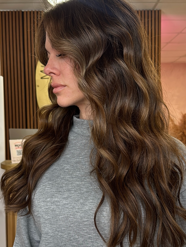 Side profile of a woman with long wavy brown hair wearing a gray top indoors.