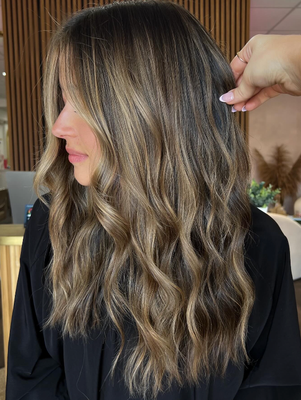 Wavy highlighted brown hair styled in loose layers, shown in profile with a hand adjusting it.