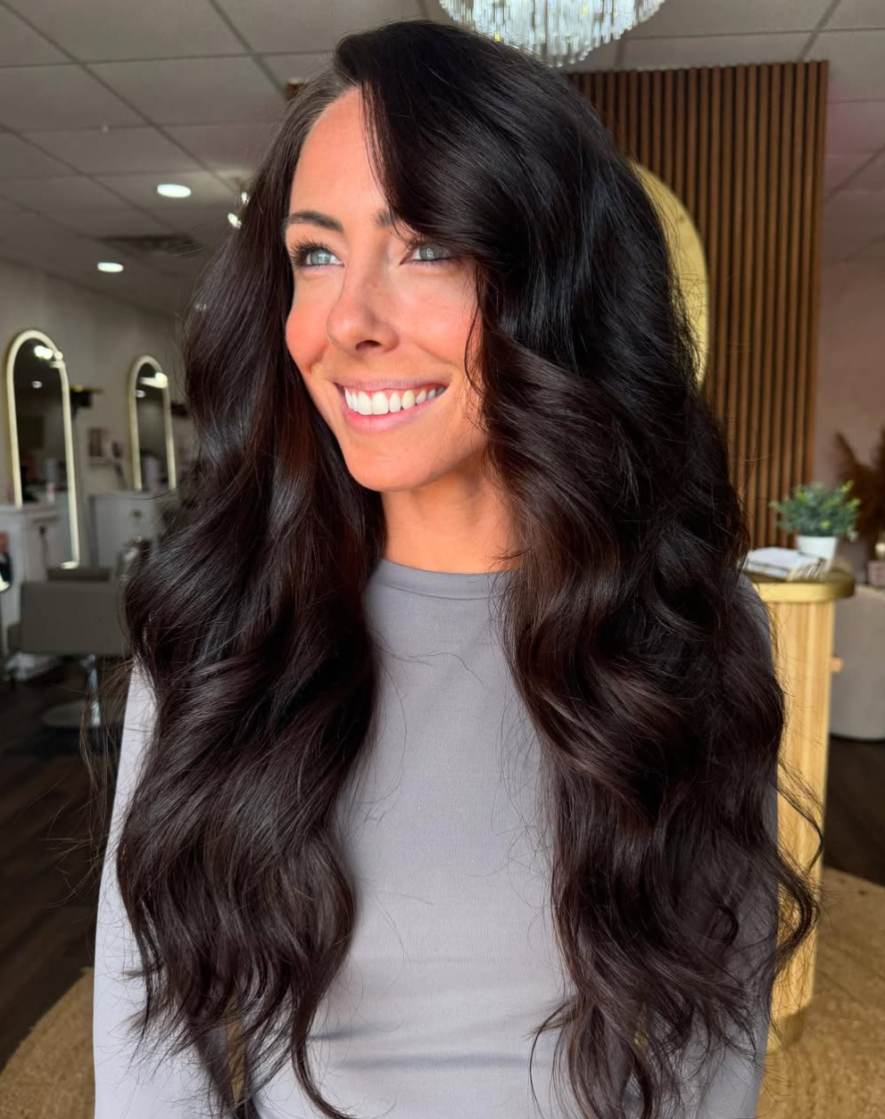Woman with long wavy dark hair smiling in a modern salon, wearing a gray top.
