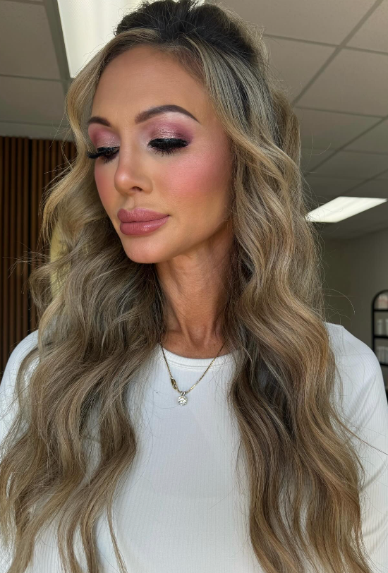 Portrait of a woman with long wavy blonde hair, pink makeup, and a white top indoors