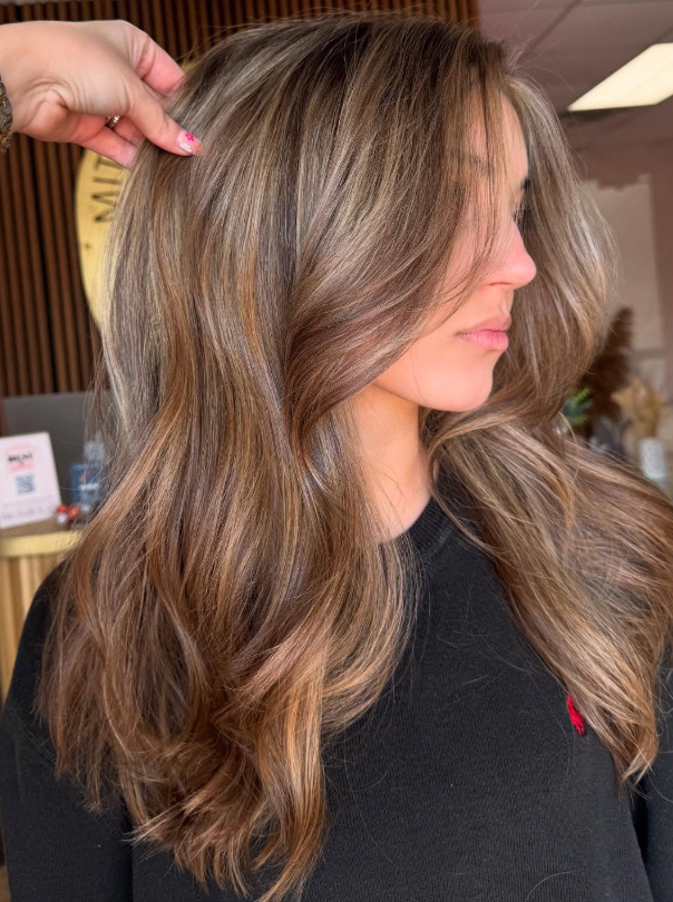 Long wavy brunette hairstyle with warm blonde highlights, styled in a salon setting