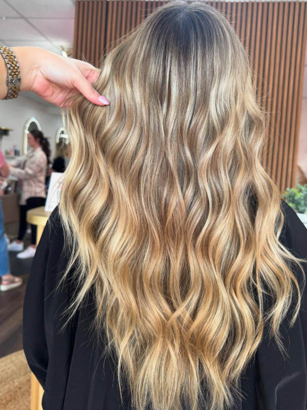 Wavy blonde balayage hairstyle with darker roots, shown from the back in a salon setting