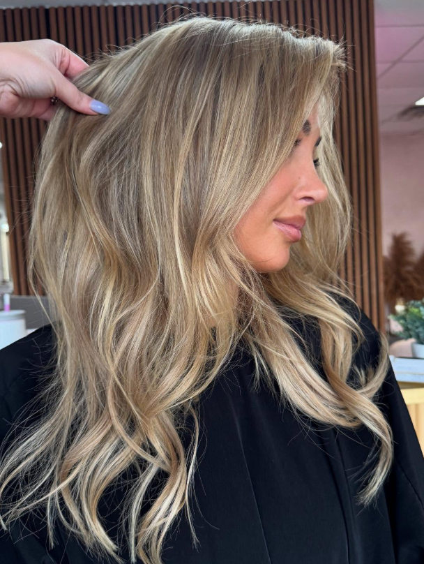 Side-profile of a woman with long blonde wavy hair being held up in a salon setting