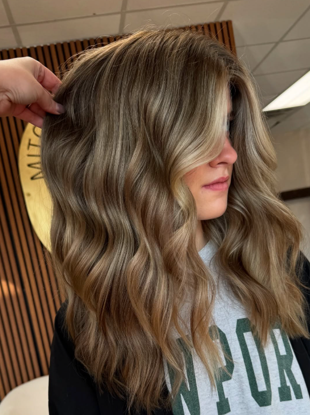 Long wavy blonde hair with dark roots, held up by a hand in an indoor setting
