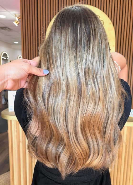 Back view of long wavy blonde hair with brunette lowlights, held by a hand in a salon.