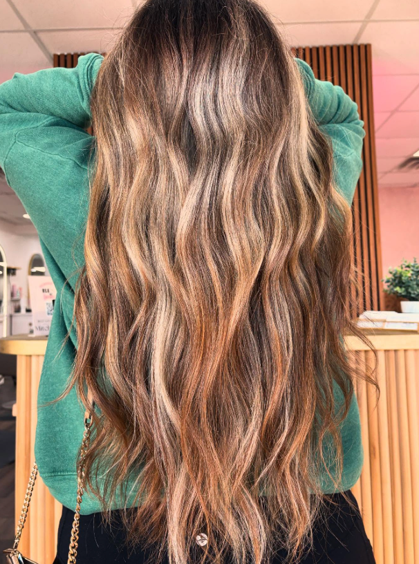 Long wavy highlighted brown hair seen from behind, with hands raised in a salon setting