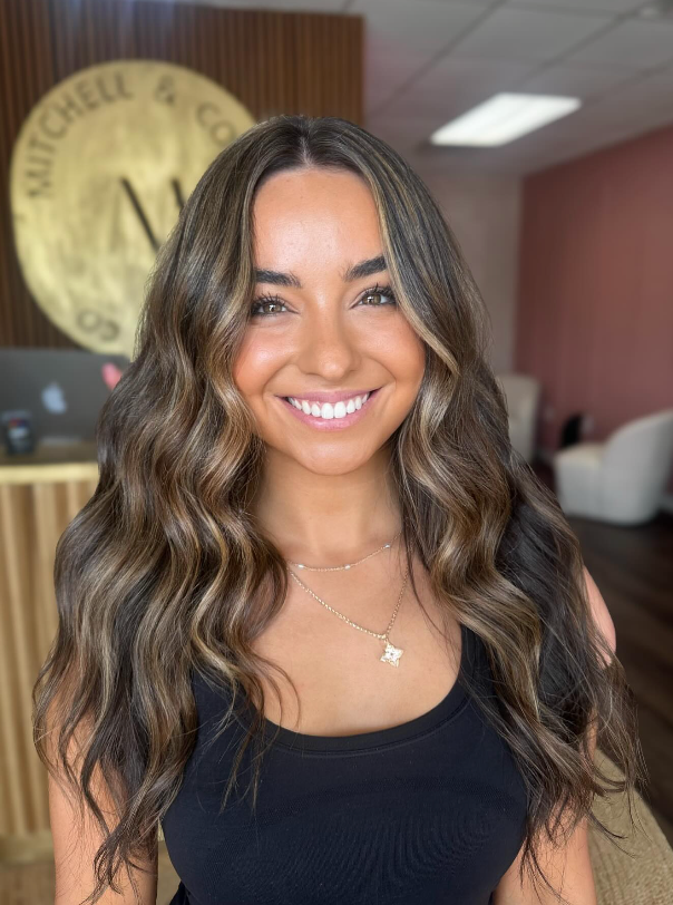 Smiling woman with wavy hair in a black top, standing indoors near a reception desk.