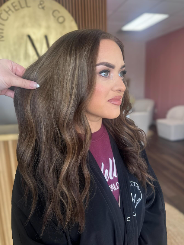 Woman with wavy brown hair in a salon, shown in profile with styled hair and makeup.