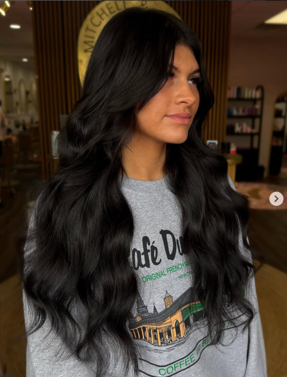 Woman with long black hair in a gray graphic sweatshirt, standing indoors and looking to the side.