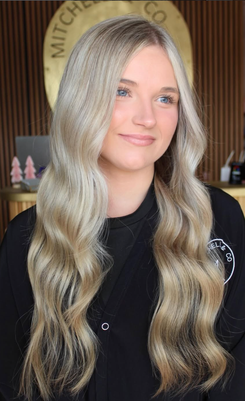 Woman with long blonde hair, wearing a black top, smiling in front of a circular sign and wooden background