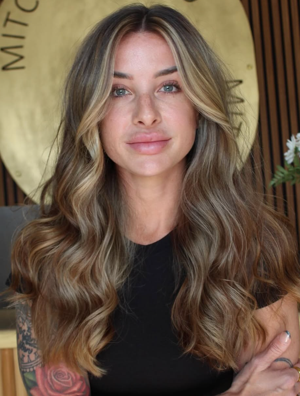 Woman with long wavy hair and black shirt, standing indoors against a wooden backdrop.