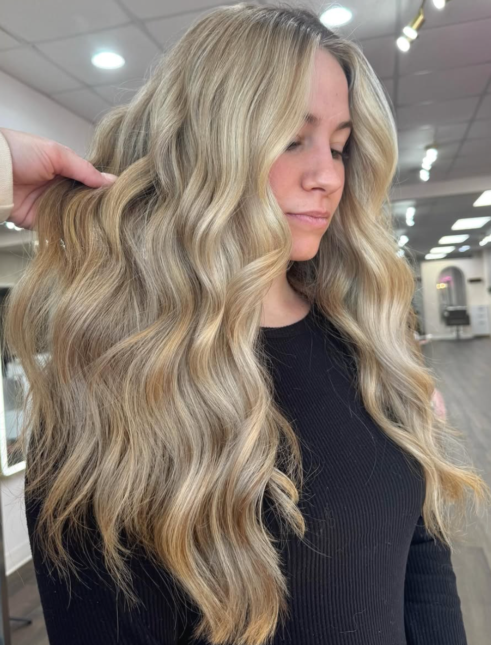 Woman with long wavy blonde hair in a salon, wearing a black top, shown in profile.