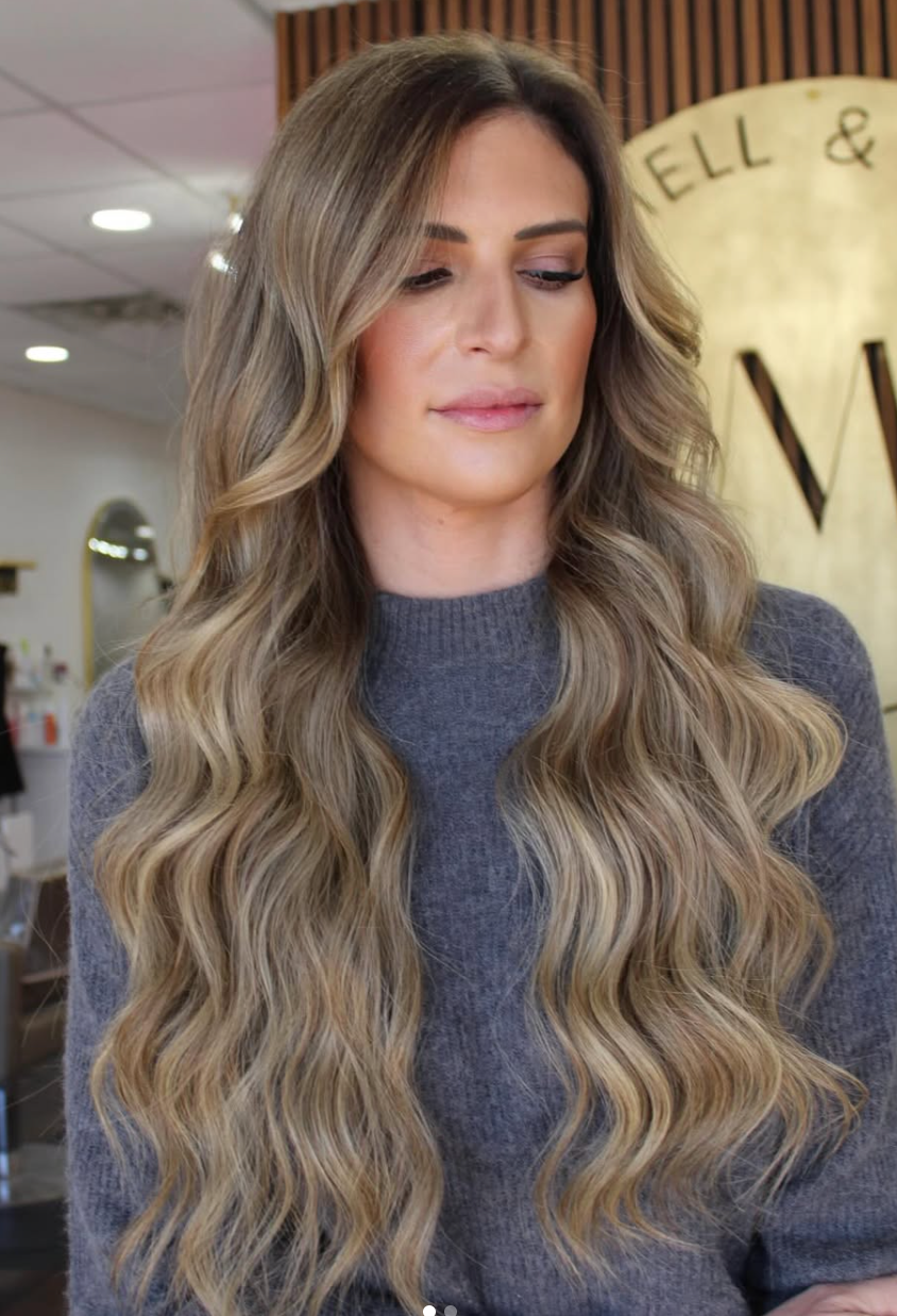 Woman with long wavy blonde hair wearing a gray sweater in a salon setting