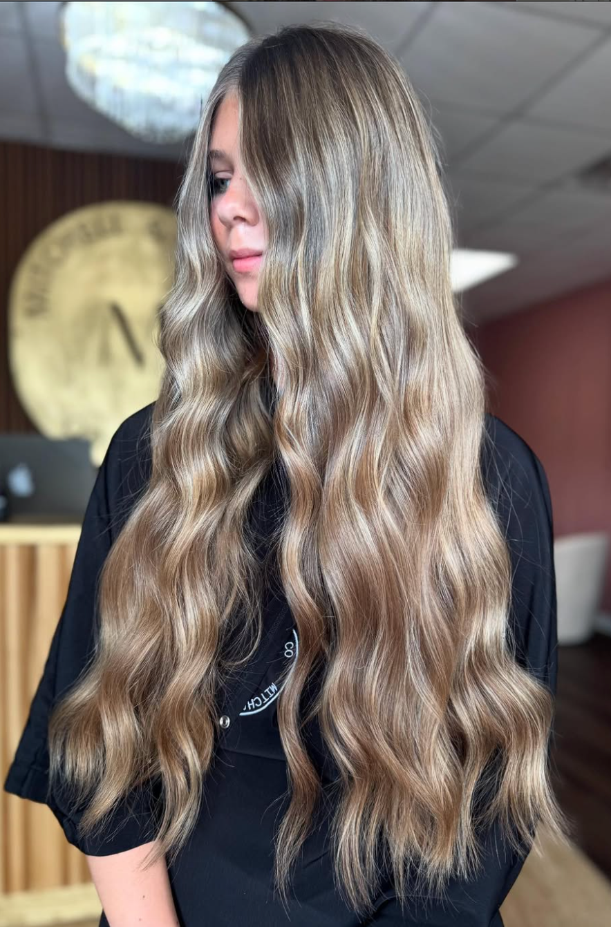 Person with long wavy blonde hair in a black top, standing indoors with a blurred background.