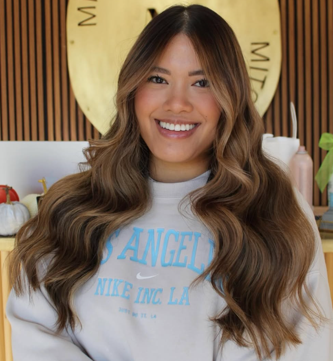 Smiling woman with long wavy hair in a gray Los Angeles sweatshirt, standing indoors