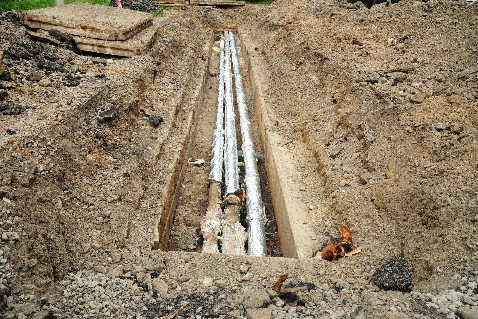 Trench with exposed metallic pipes in the ground, likely for utilities. Surrounding dirt and debris.