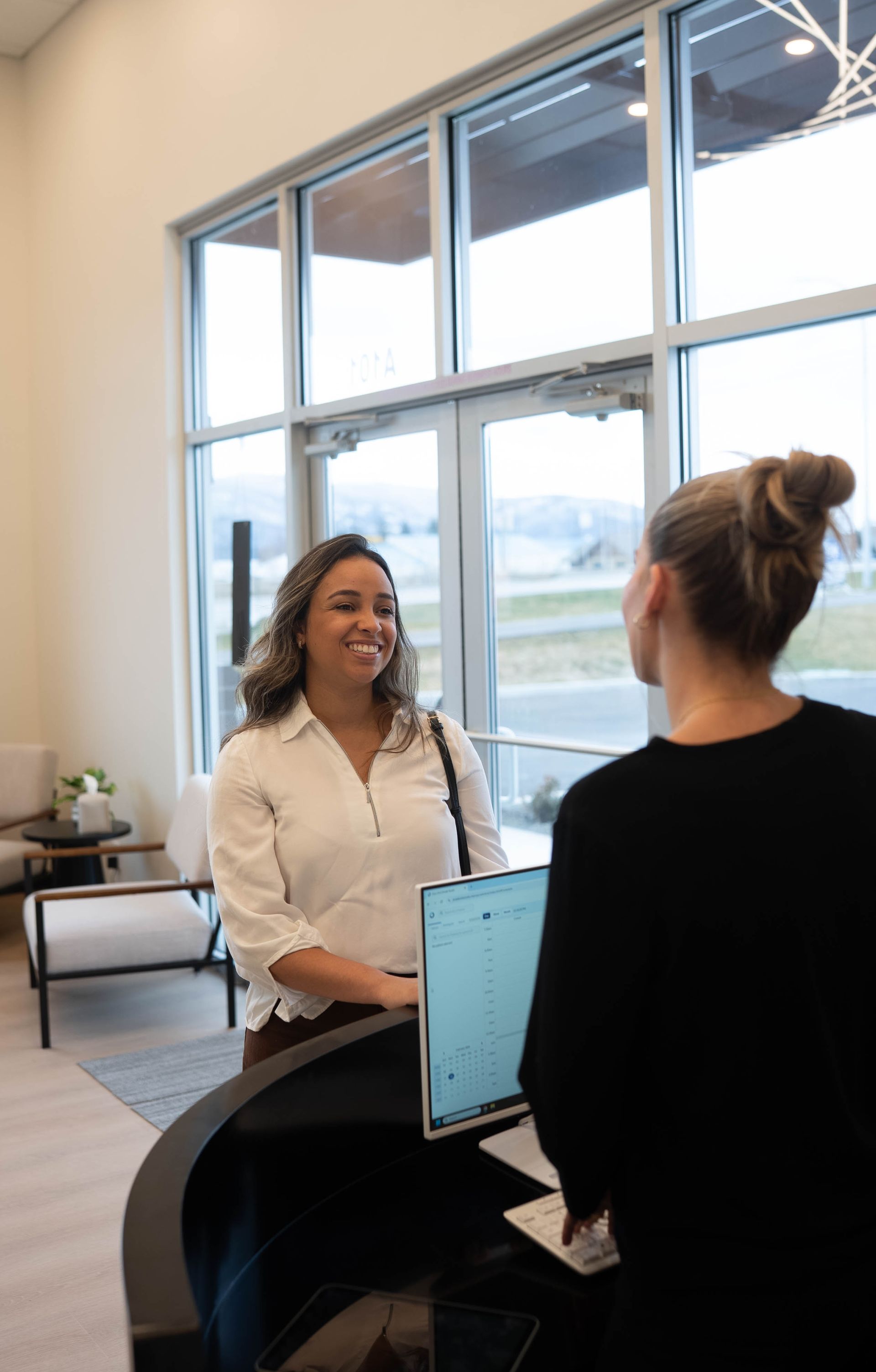 Two people chatting in an office lobby beside large windows and a laptop.