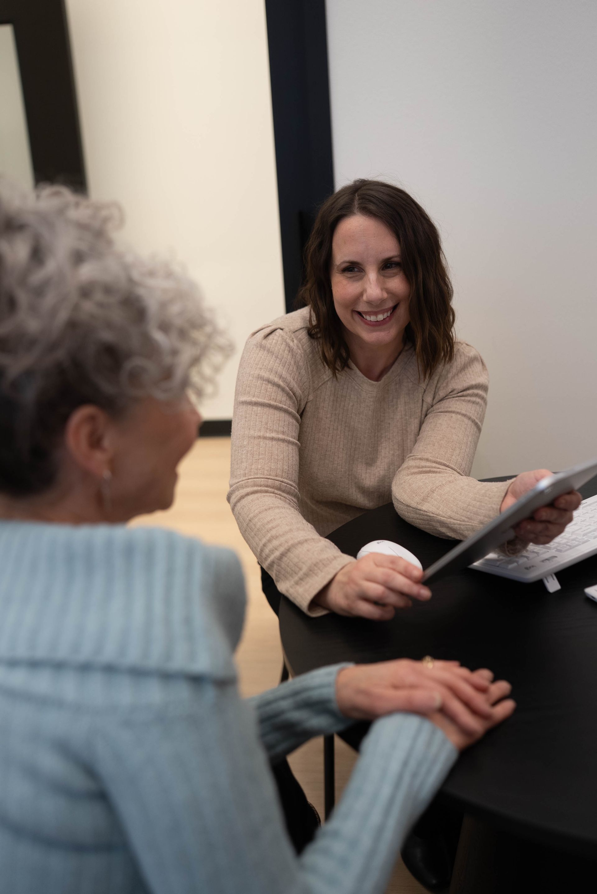 Two women talking at a table, one holding a tablet and smiling in a office setting