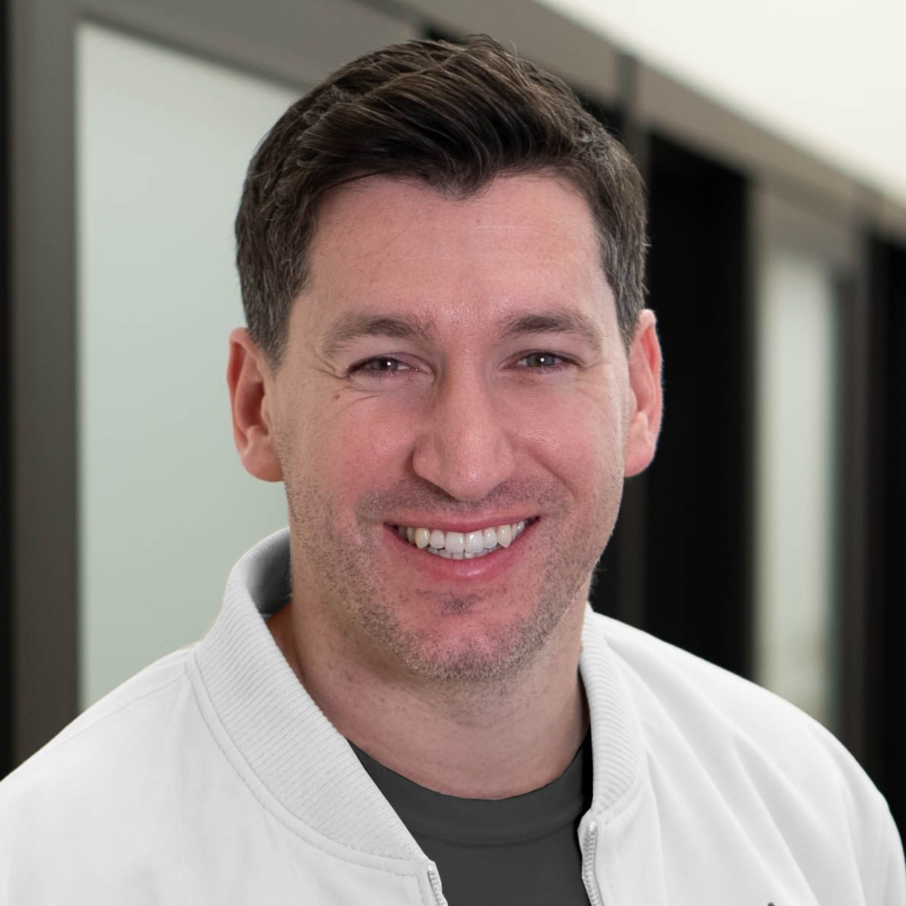 Smiling person in a white jacket, standing indoors against a blurred background