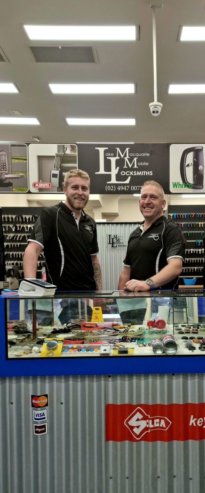 Two Men Behind a Counter in a Store, Both Wearing Black Shirts — Lake Macquarie Mobile Locksmiths in Belmont, NSW