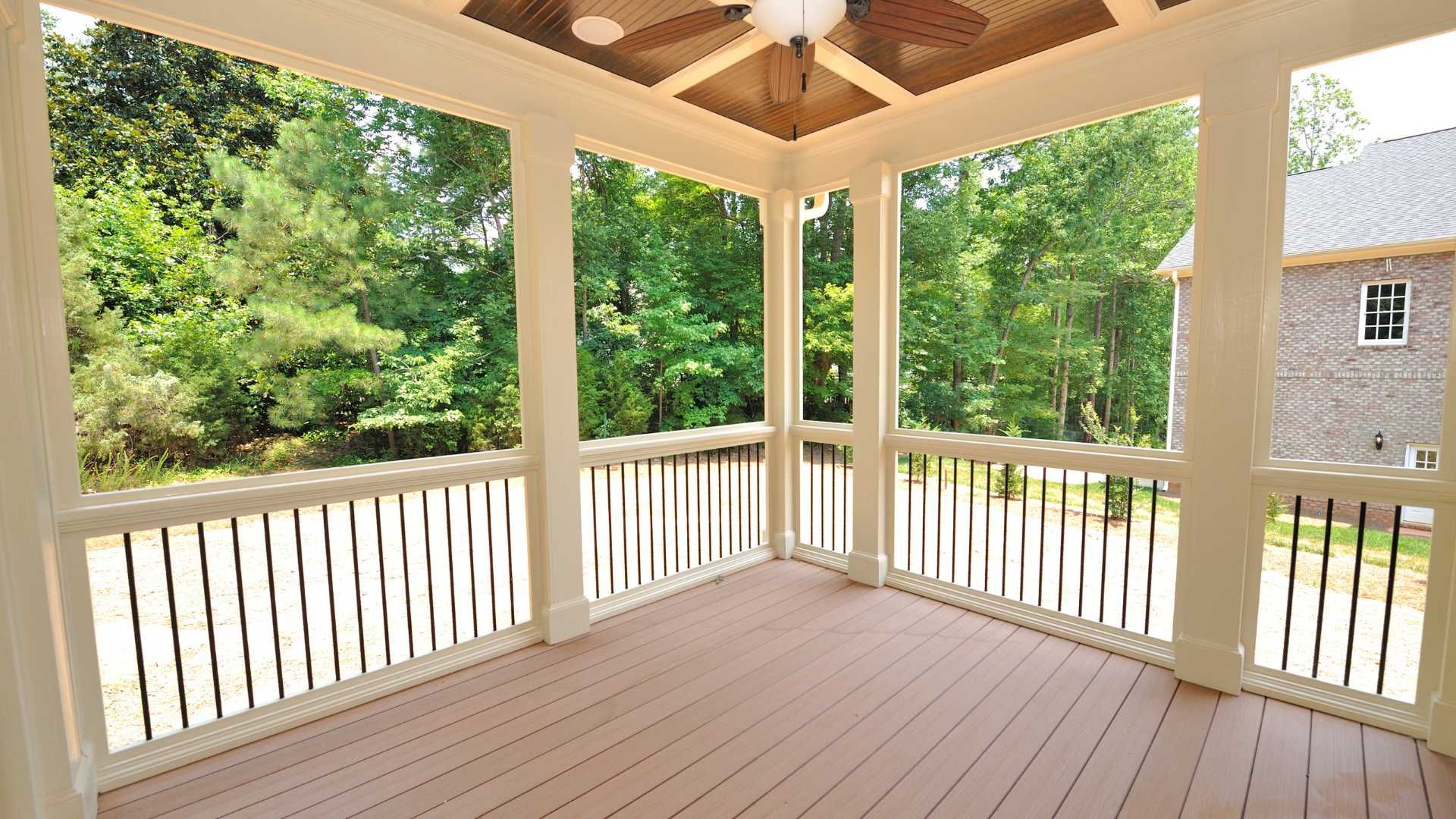 Covered wooden porch with white railings and a vaulted ceiling, overlooking green trees and a neighboring house.