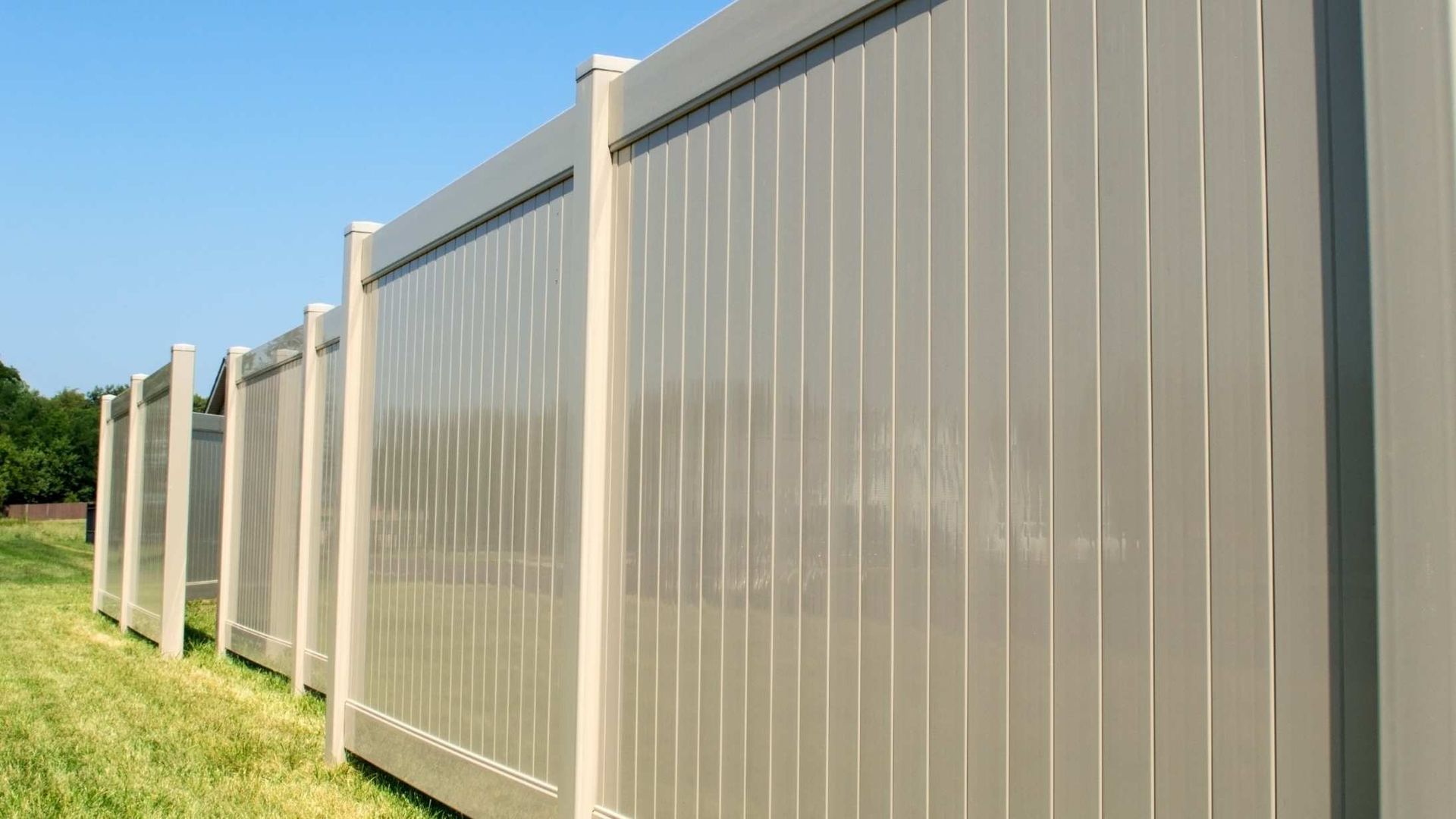 Tall beige metal privacy fence running along a grassy roadside under a clear blue sky