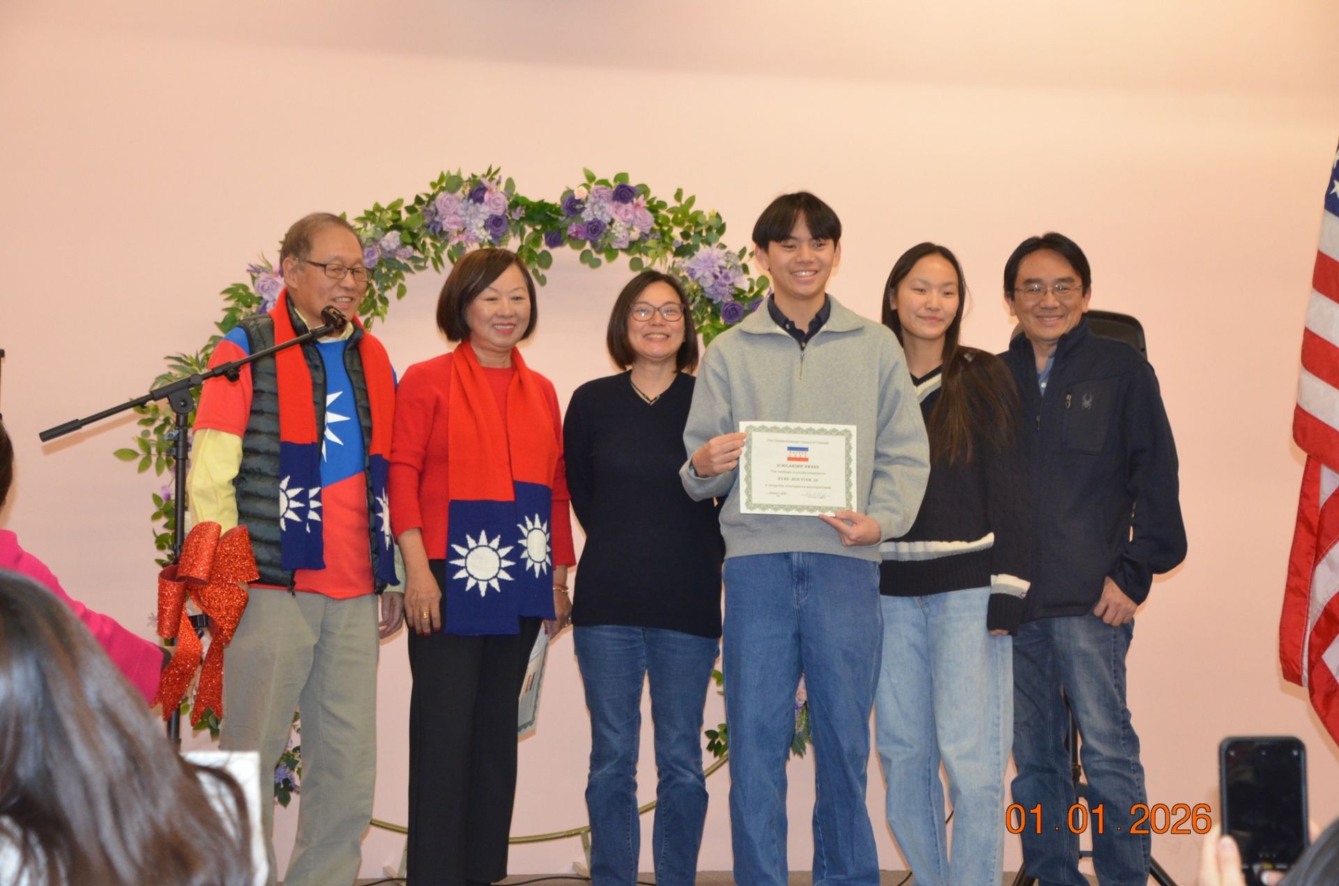 Six people on stage posing for a certificate presentation under a floral arch.