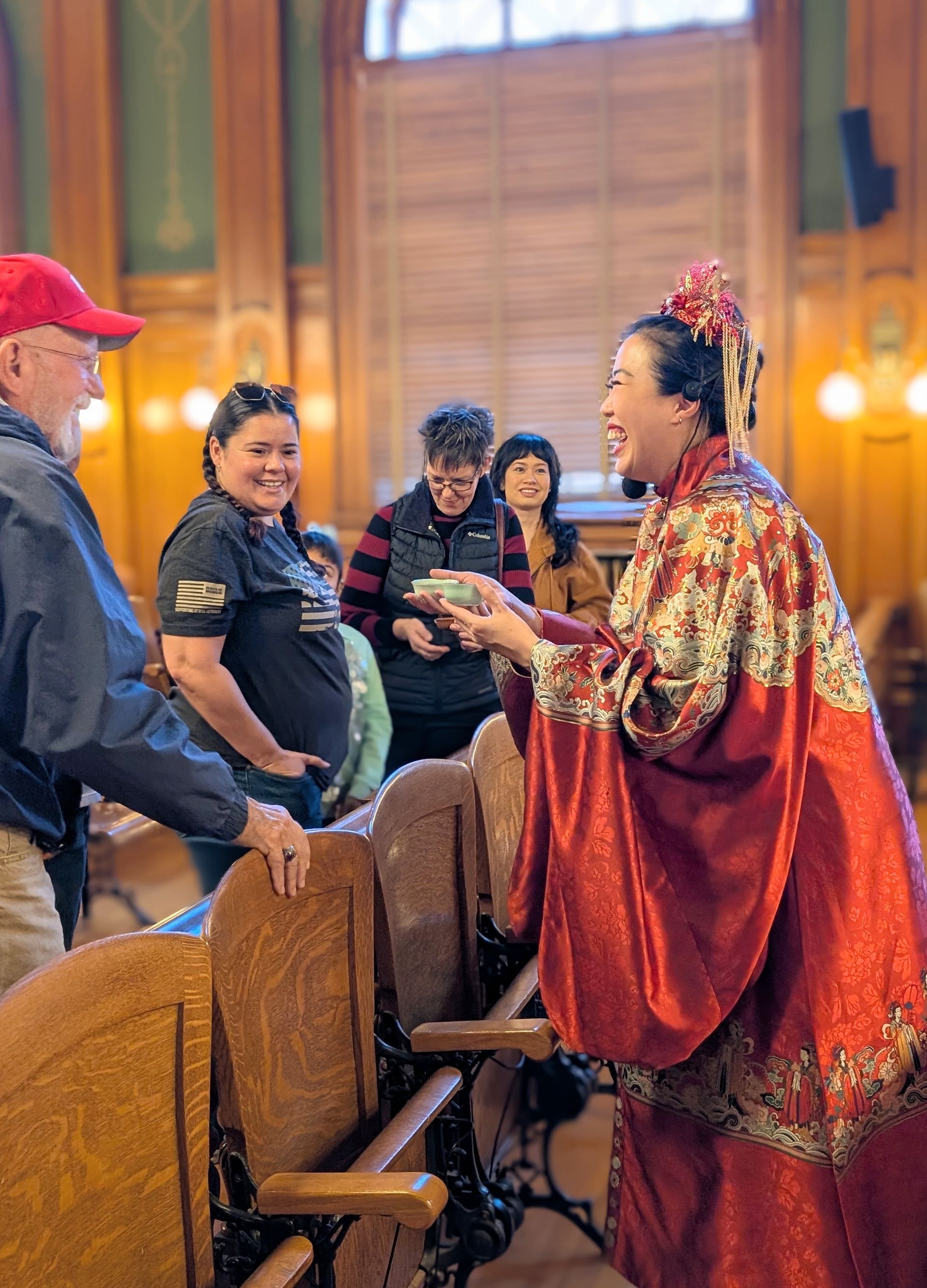 A person in a traditional red brocade robe smiles while offering a small item to a group in a wood-paneled room.