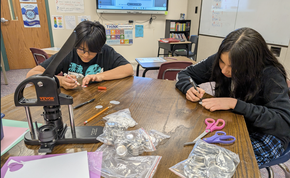 Two students work at a table with a button-making machine and craft supplies in a classroom.