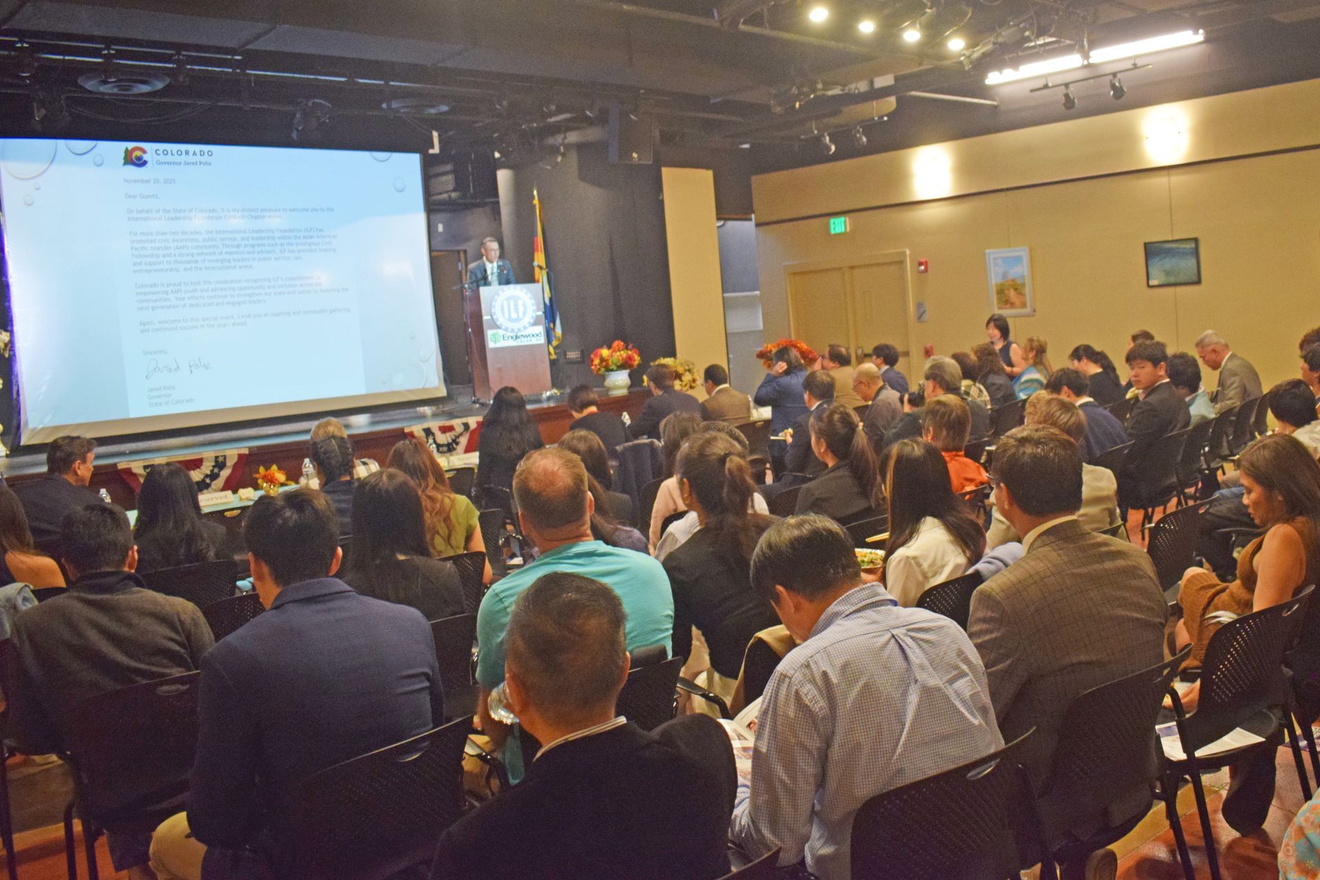 A speaker presents on a screen to a crowded audience in a conference hall.