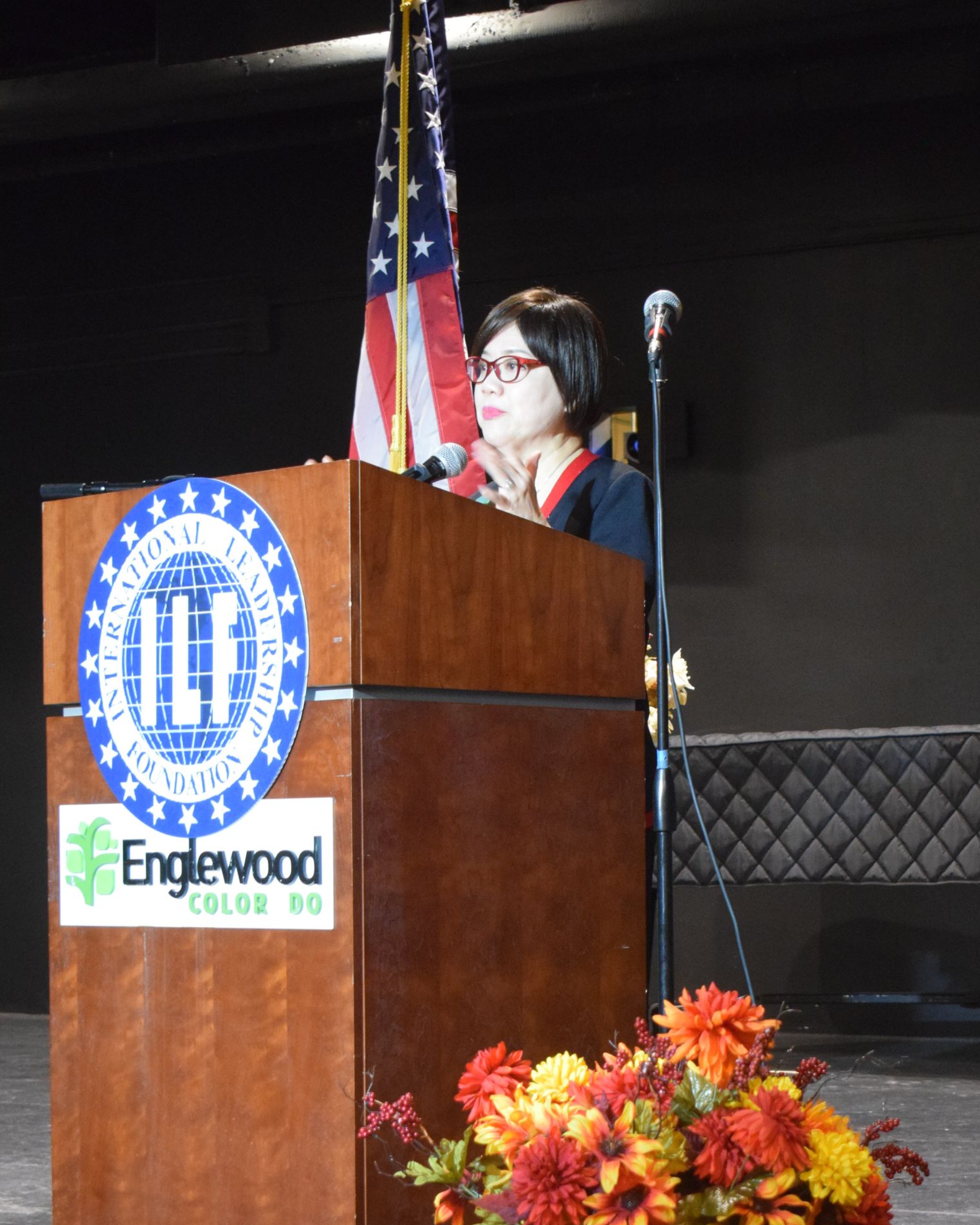 Woman speaking at a podium with a city seal and US flag in the background. Autumn flower arrangement in front.