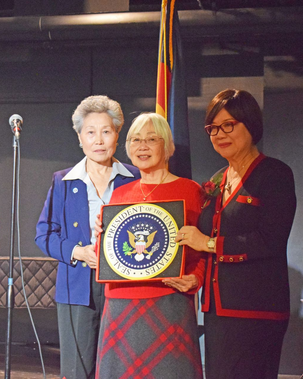 Three women holding a plaque with the U.S. presidential seal. Indoors, one woman in a red sweater, others in blue and black.