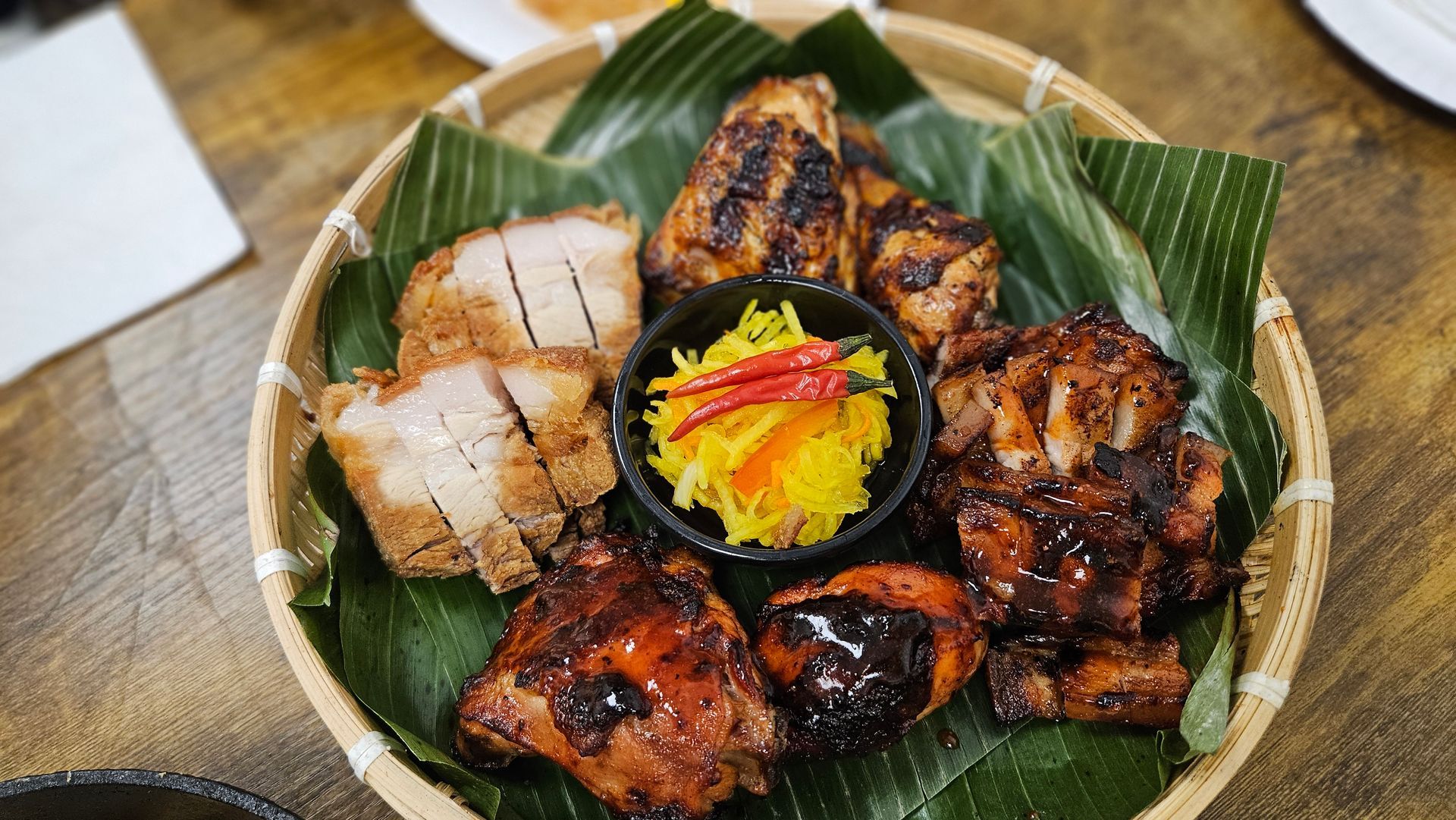 Grilled meats platter with pickled vegetables on a woven basket lined with green leaves.