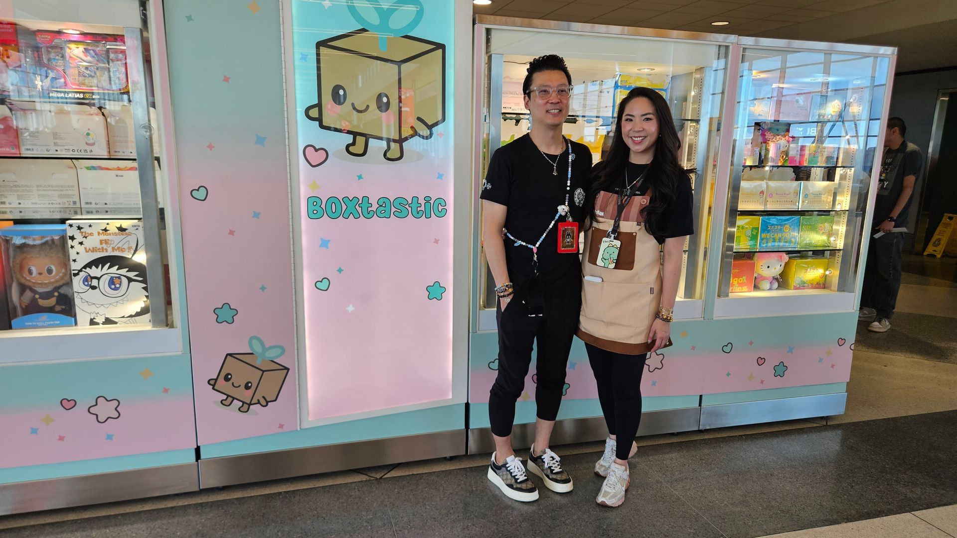 Two people stand smiling in front of a brightly colored, pastel claw machine game kiosk in a shopping area.