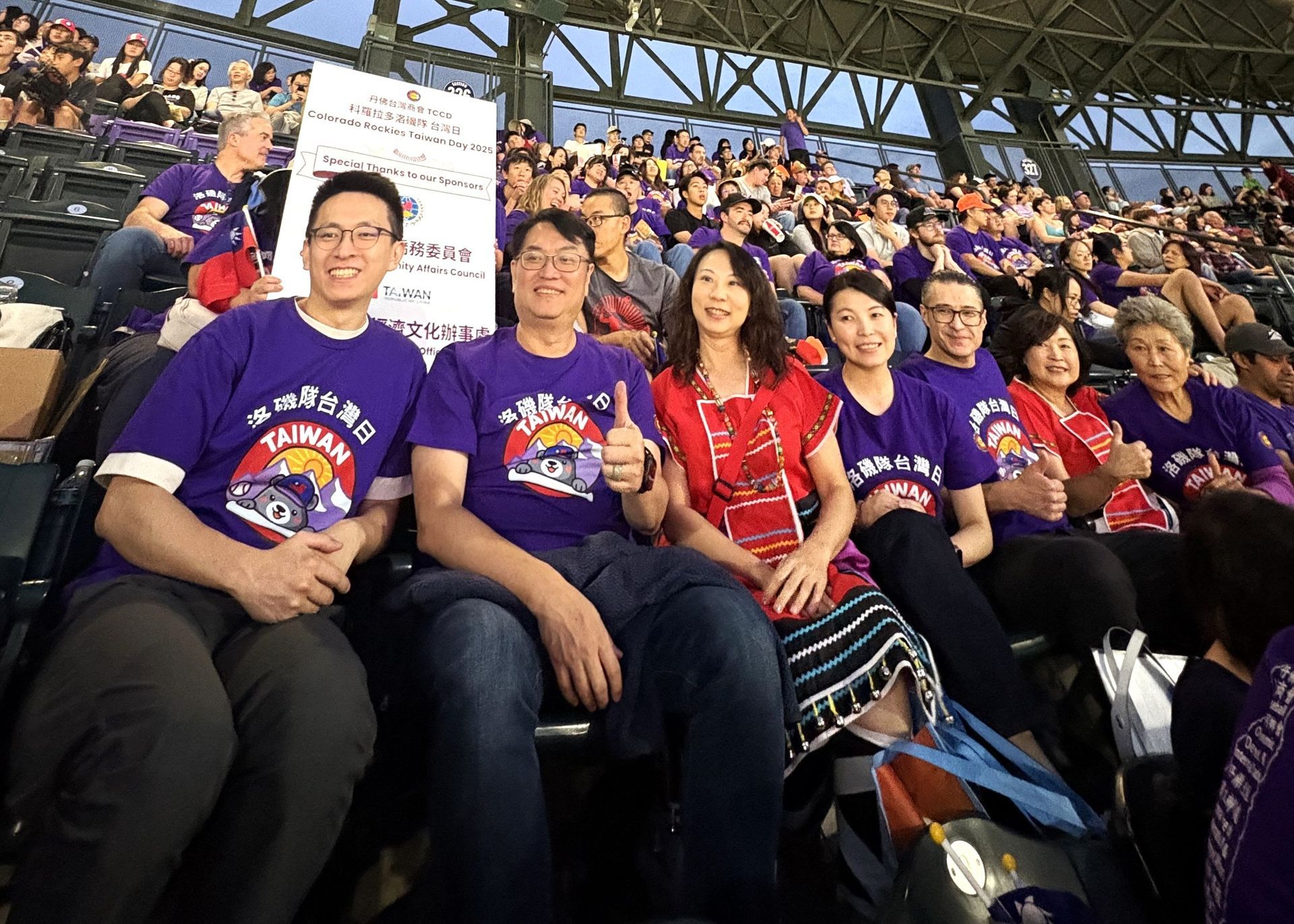 Group of people in purple shirts at a stadium, some giving thumbs up. Fans in stands above.