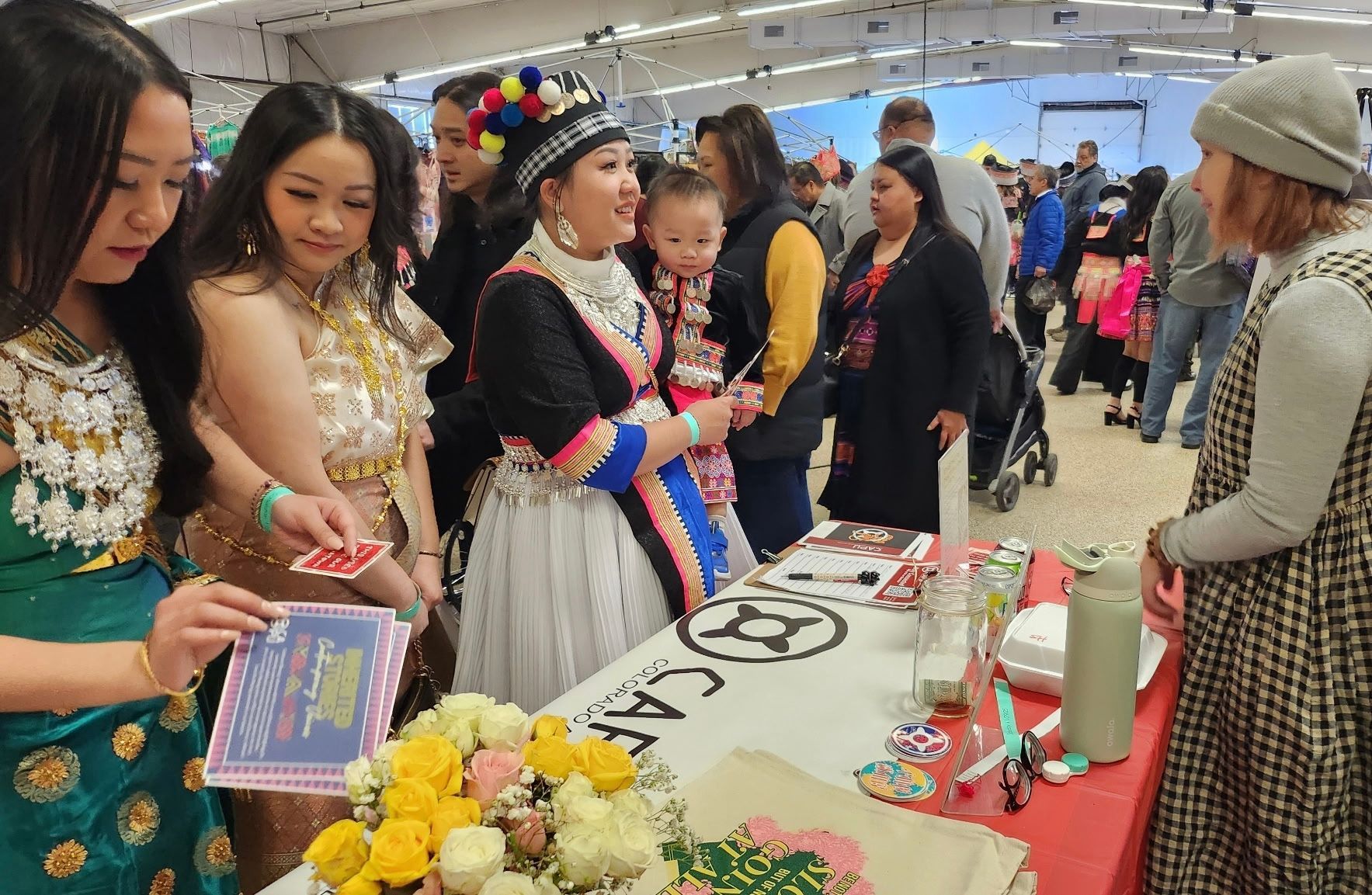 People at a cultural event, women in traditional dress, interacting with attendees at a booth indoors.