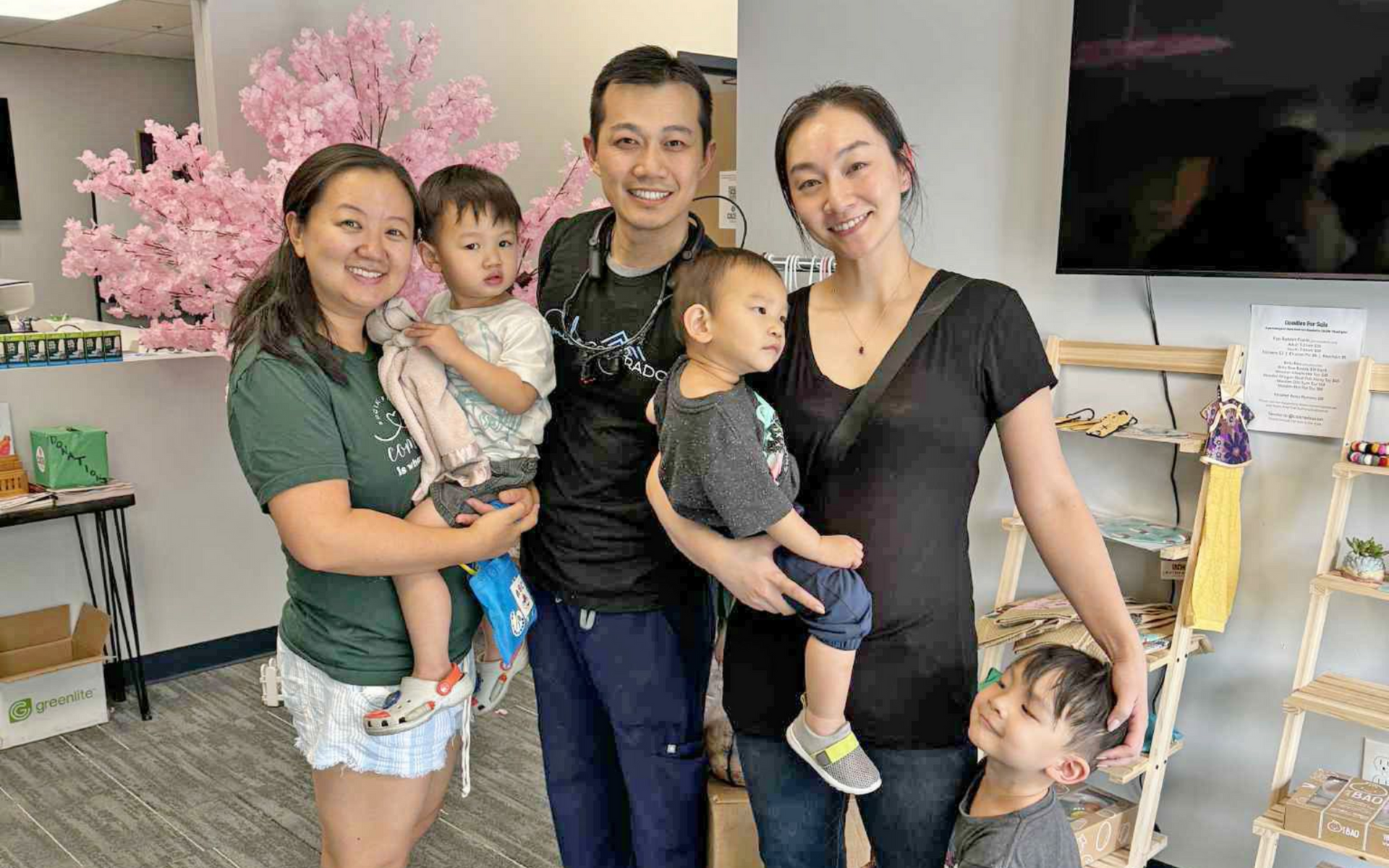 A group of five people, two adults holding toddlers and another child smiling, by pink decorations.