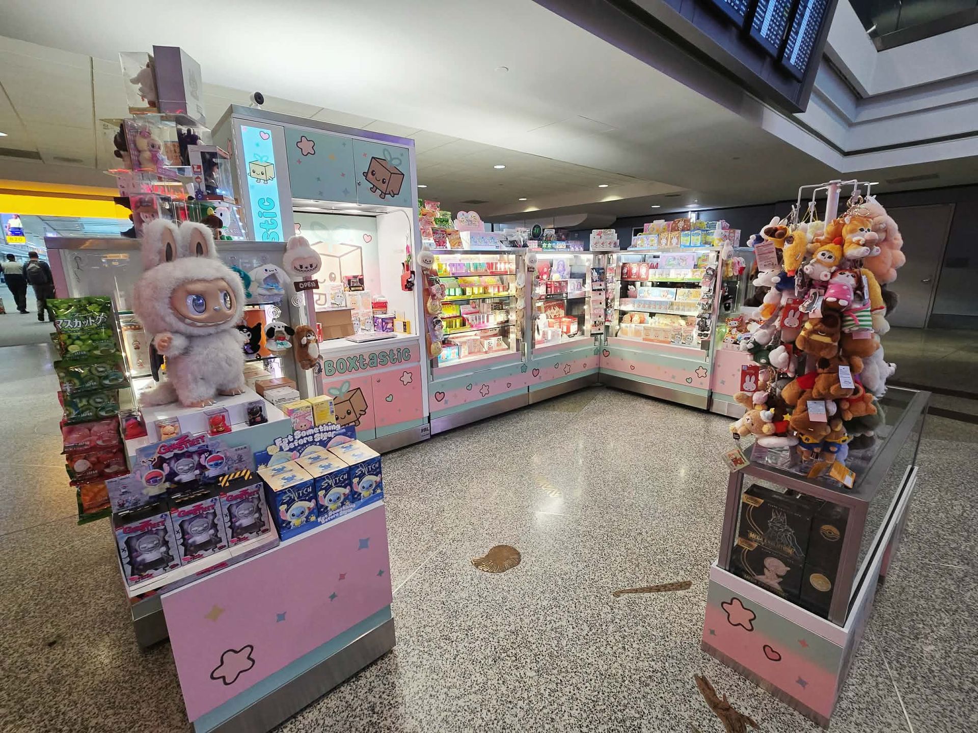 A colorful pop-up kiosk in a bright indoor shopping area, displaying plush toys, character goods, and various snacks.