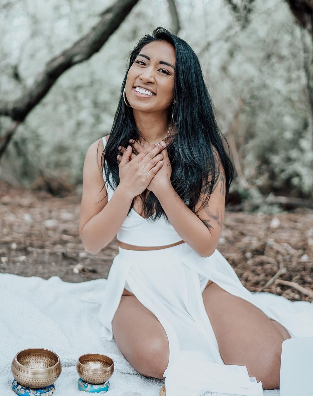 Christina Ifurung in white outfit outdoors with hands on chest, smiling near singing bowls.
