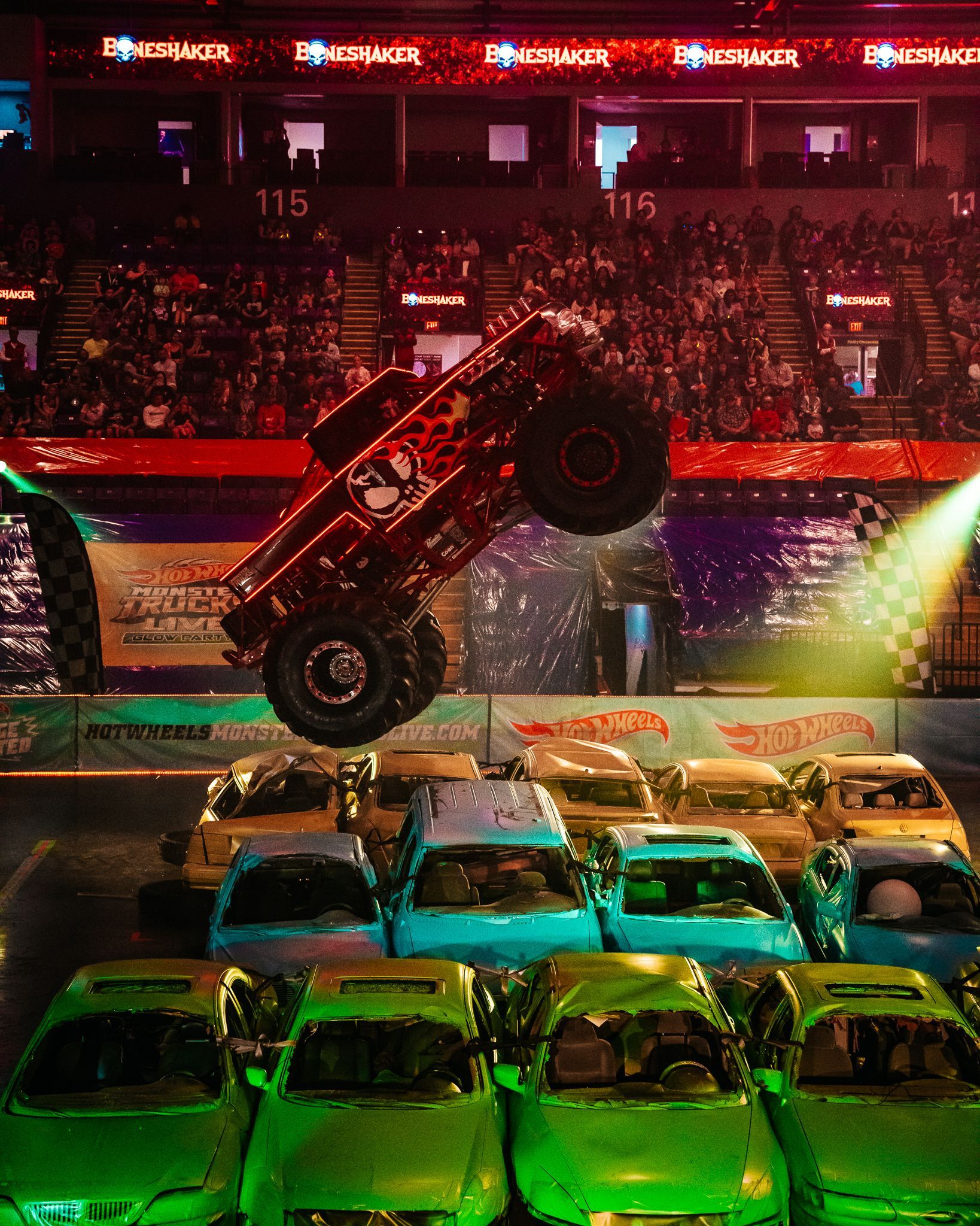 Monster truck in mid-air, performing a stunt over crushed cars at an indoor arena.