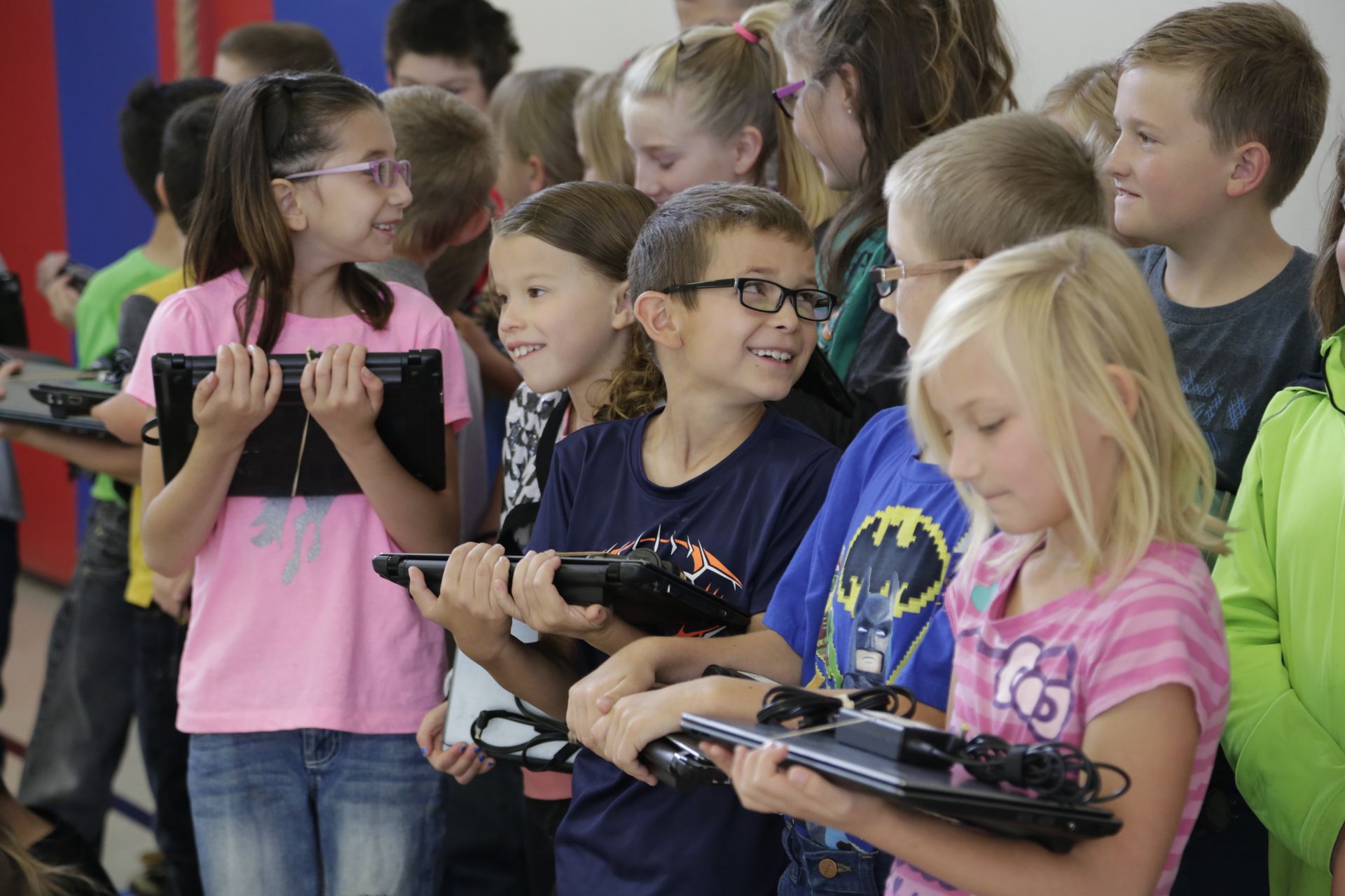 School children holding laptops, looking at a device, and smiling. Brightly colored room, natural light.