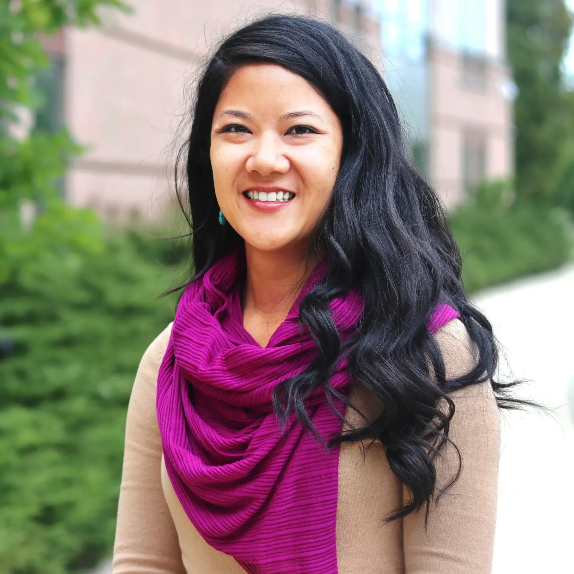 Daranee Teng with a purple scarf smiles, set outdoors near greenery and a building.