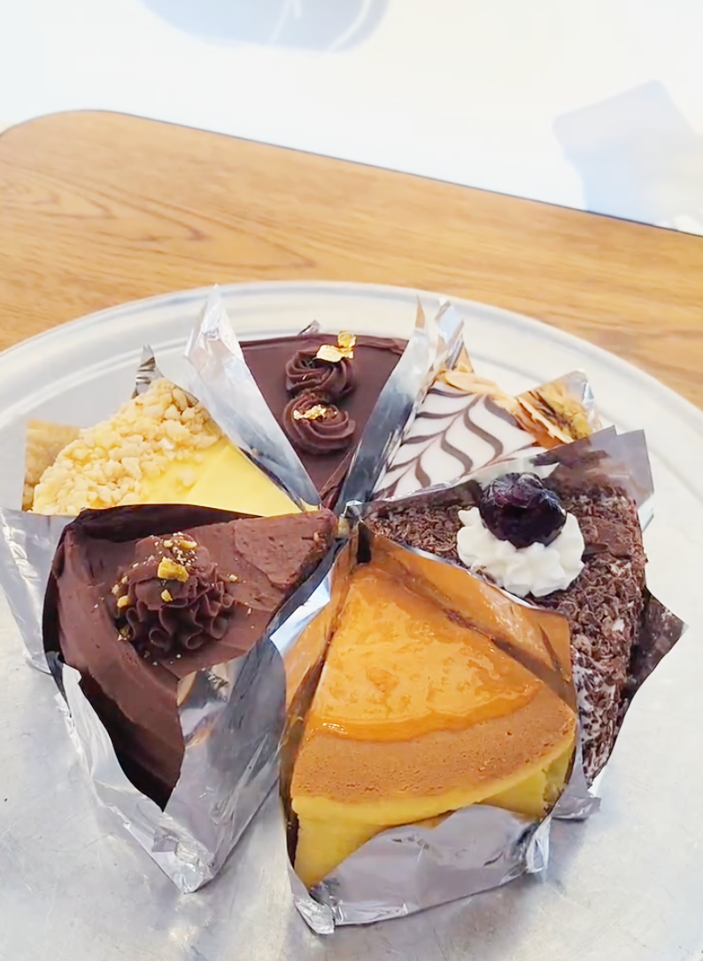 Assortment of cake slices on a silver platter, each wrapped in foil. Flavors vary, with chocolate and fruit.