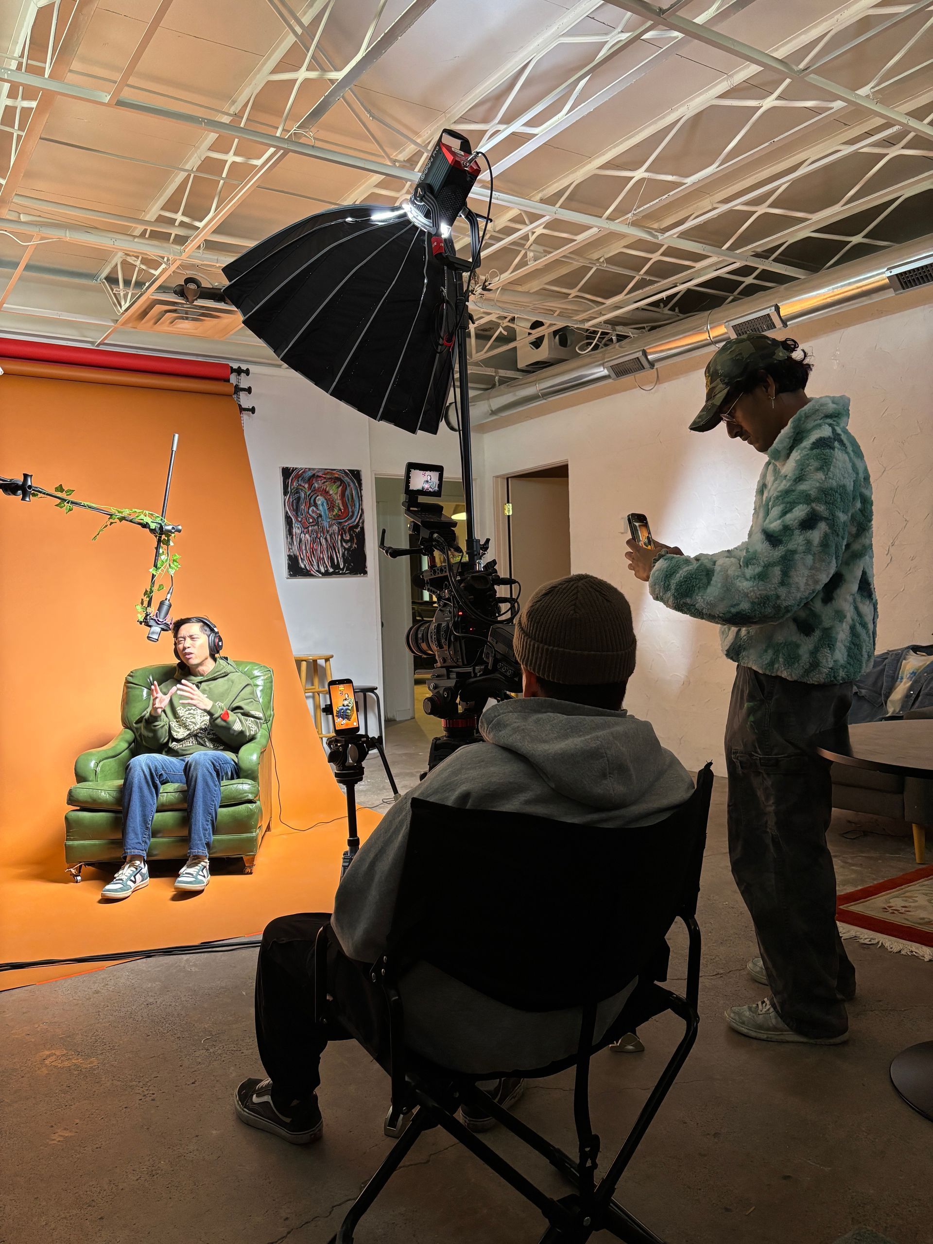 Photoshoot in studio: person on orange set, photographer, assistant, and overhead softbox light