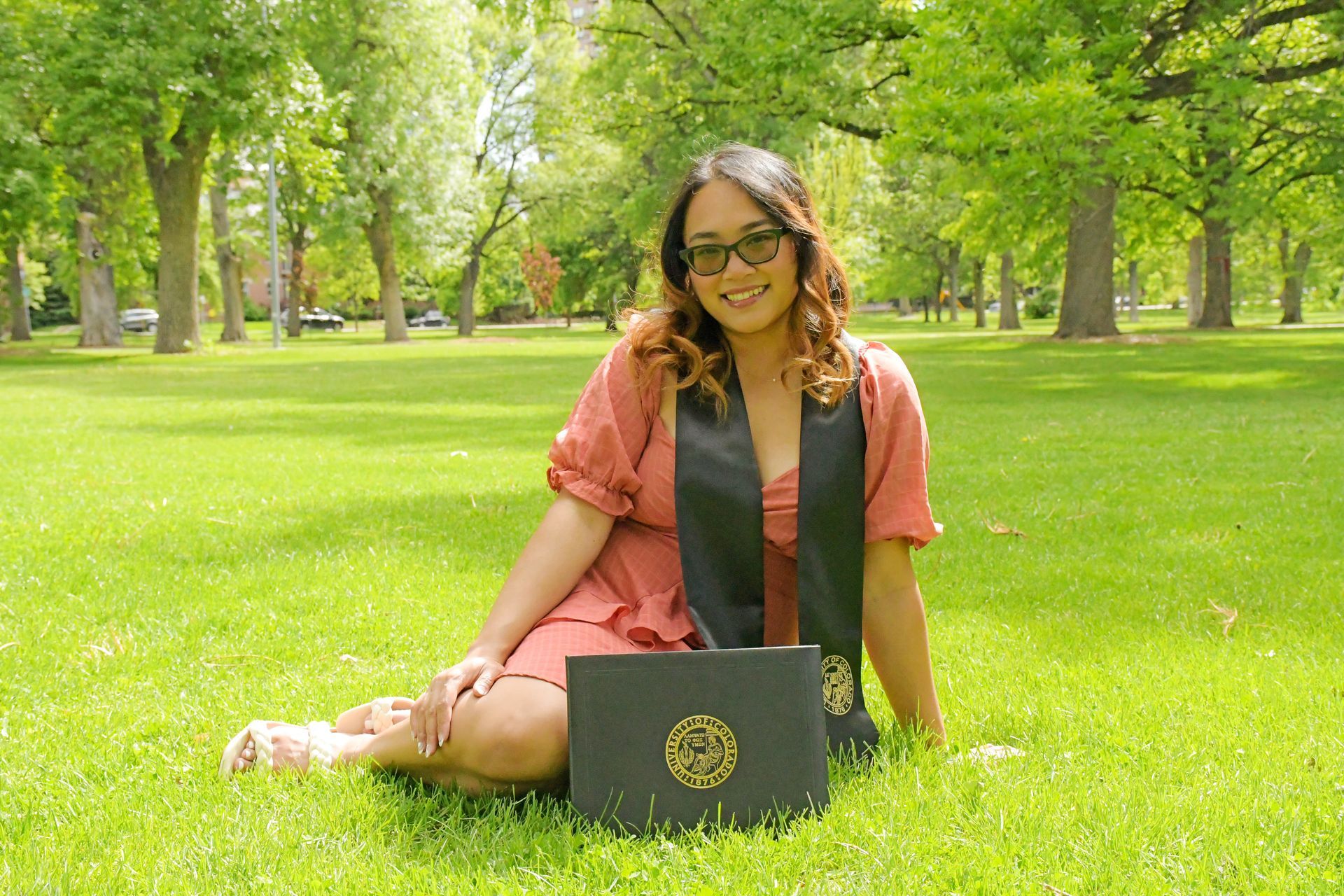 Woman in graduation gown sitting in grass with diploma, smiling. Sunny outdoor setting.