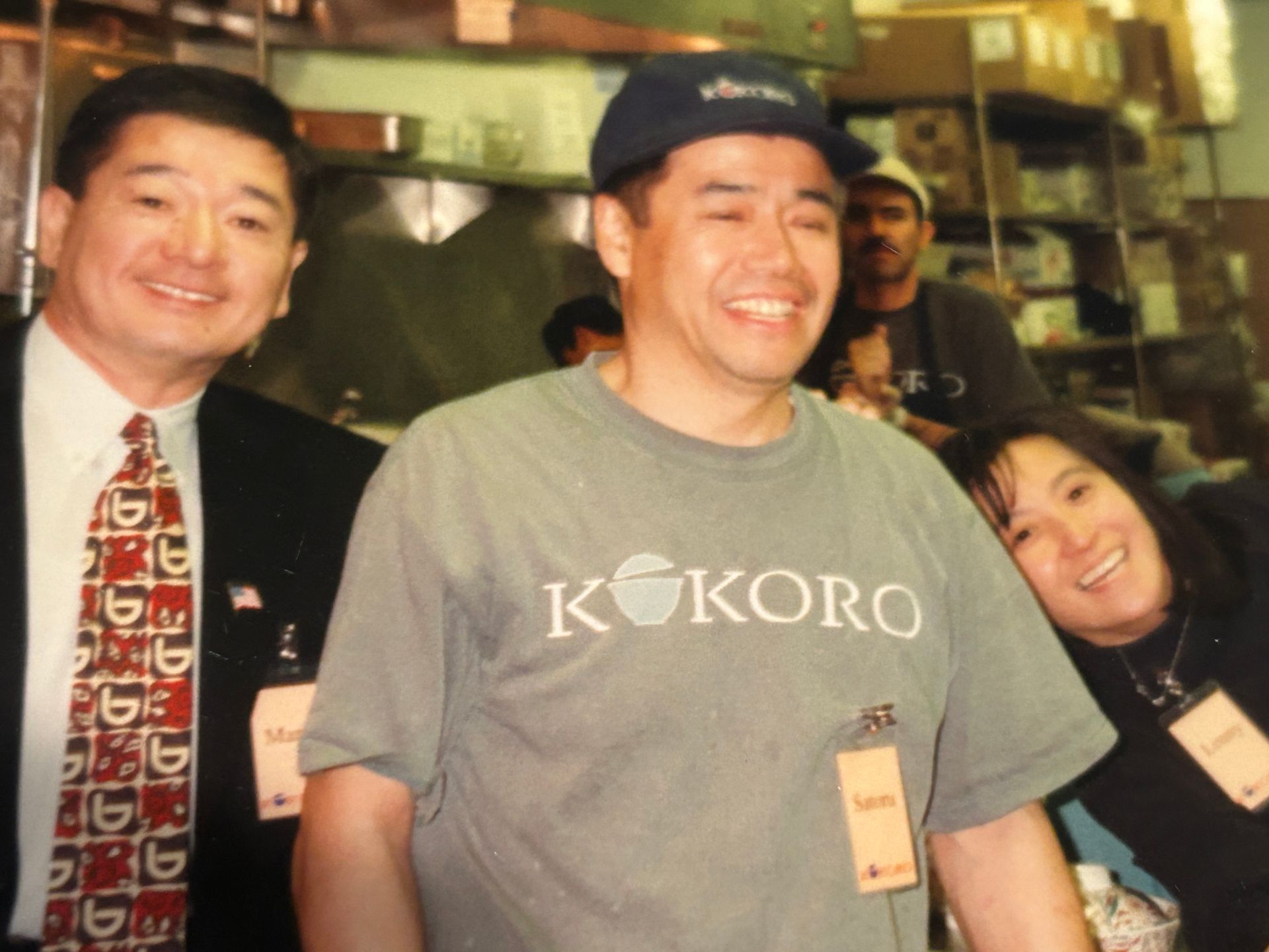 Three smiling people posing indoors, with one wearing a gray “Kokoro” shirt and cap.