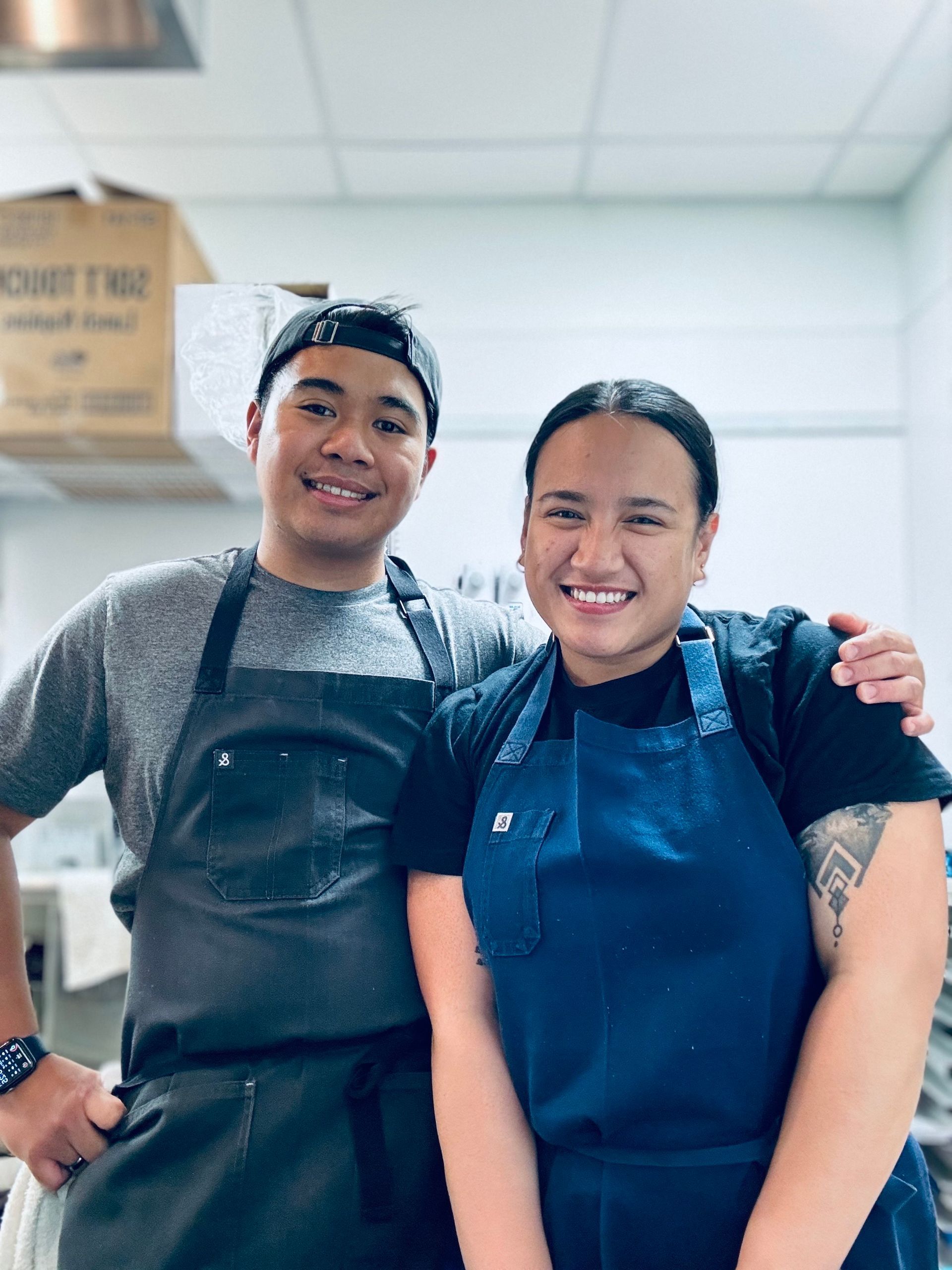 Two people in aprons smiling, standing in a kitchen.