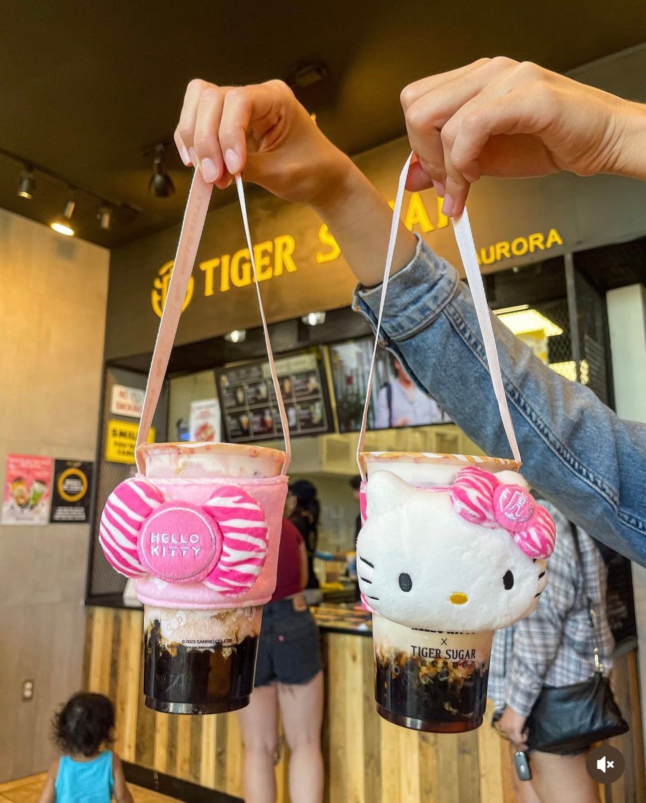 Two drinks in pink and Hello Kitty cup holders are held up by hands inside a boba tea shop.