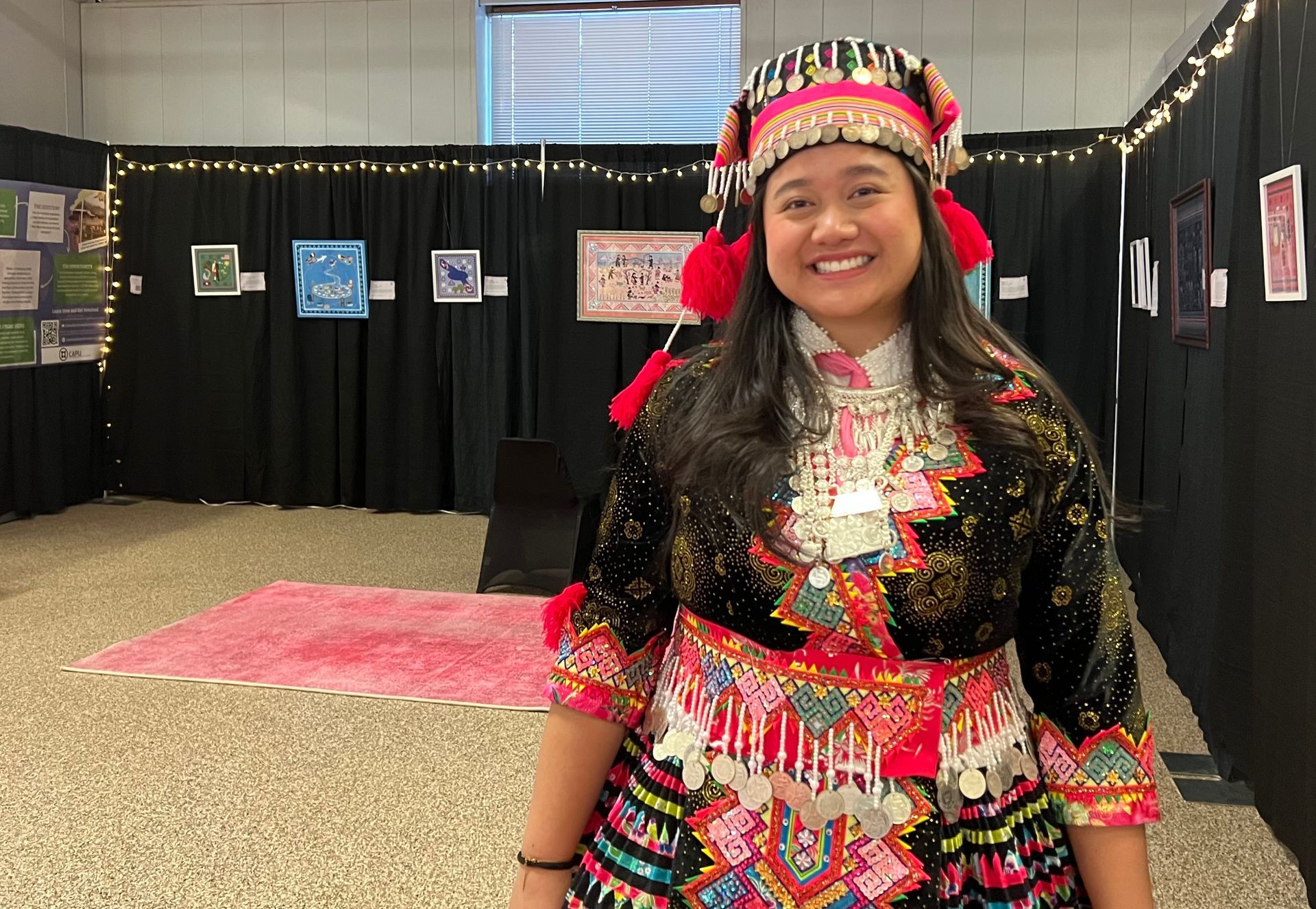 Person in traditional Hmong clothing smiles in an art exhibit, standing in front of black curtains and artwork.