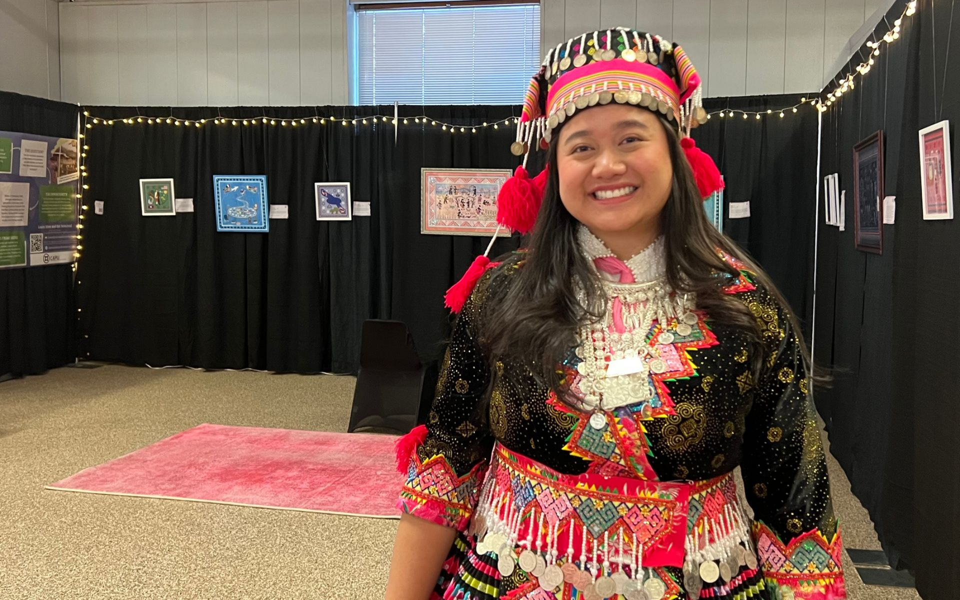 Woman in traditional clothing smiles, posing in front of her art display at an event.